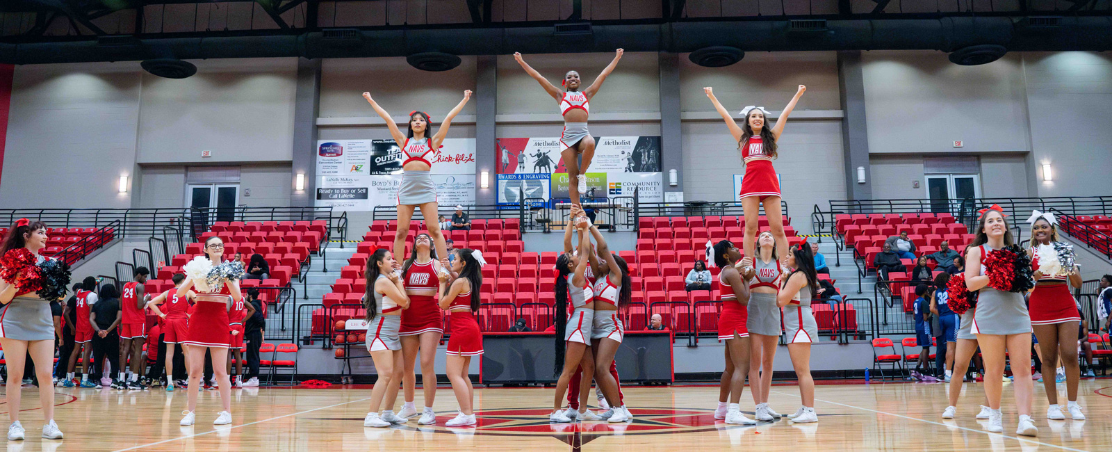 The cheer team at a basketball game in the arena. Three small pyramids and other cheerleaders with pompons.